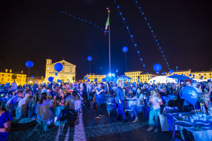 Evento serale in Piazza Grande: tavolate affollate sotto palloncini blu e luci, con il Duomo illuminato sullo sfondo.