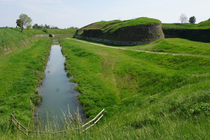 Un bastione di Palmanova: alte mura in pietra coperte d'erba e, in primo piano, un fossato con acqua tra due prati verdi.