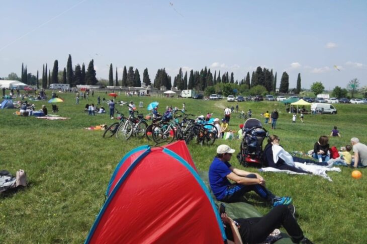 Pasquetta sui bastioni: persone sedute sul prato con biciclette e tende, aquiloni nel cielo e cipressi sullo sfondo.
