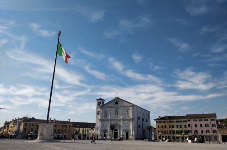 Piazza Grande a Palmanova: una grande bandiera italiana su un alto pennone e, sullo sfondo, il Duomo bianco con il campanile.