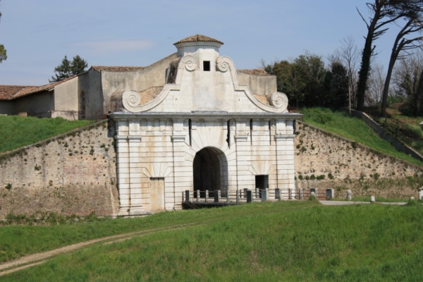 Porta Aquileia: facciata in pietra chiara con arco centrale, accanto alle mura erbose.