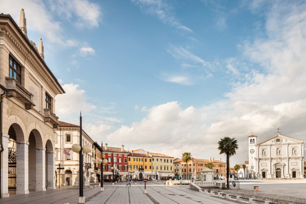Veduta parziale della piazza centrale della città di palmanova dove sono presenti a sinistra l'edificio del comune e destra si vede il Duomo del Santissimo Redentore, in centro all'immagine sono presenti degli edifici clasici e colorati della città.