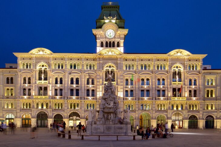 Municipio di Trieste illuminato di sera, con la torre dell’orologio al centro.